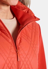 Frau trägt eine wattierte rote Weste mit Reißverschluss über einem beigen Stricktop, das den Kragen, den Reißverschluss und die strukturierten Stoffdetails zeigt.