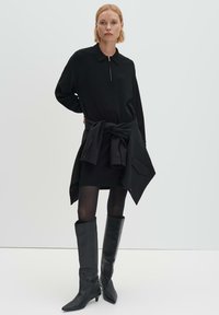 Abito nero a maniche lunghe con colletto con zip, indossato con uno strato esterno nero annodato e abbinato a stivali di pelle neri al ginocchio.