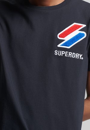 Osoba nosząca czarną koszulkę z logo Superdry w kolorach czerwonym, białym i niebieskim na lewej górnej części klatki piersiowej.