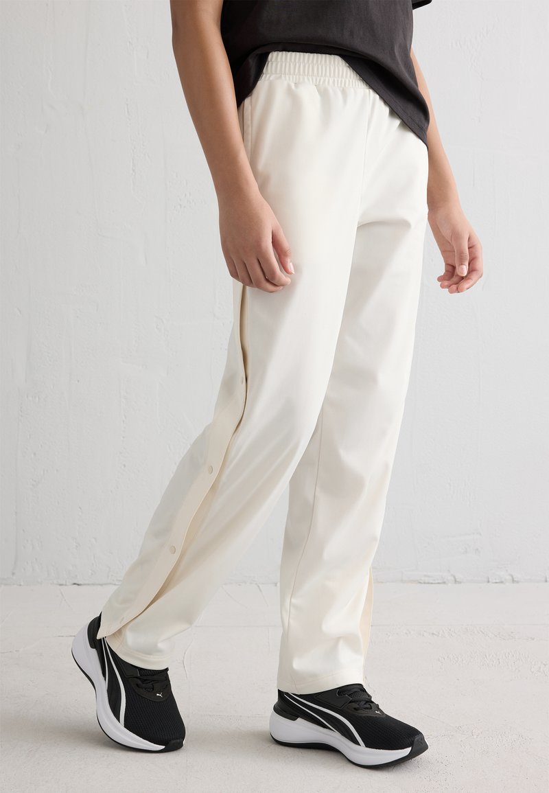 Pantalones blancos ligeros con una cinturilla elástica y botones de presión en los lados; combinados con zapatillas negras que tienen una suela blanca y gris.