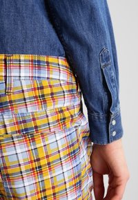 Jeansskjorta med en blå kropp och knappärmar, ihopparad med flerfärgade byxor i rutiga mönster med gult, rött, blått och svart. Texturerad tyg.