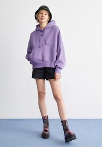 Sweatshirt à capuche violet avec poche avant, short noir court et bottes lacées noires et violettes brillantes sur un sol bleu.