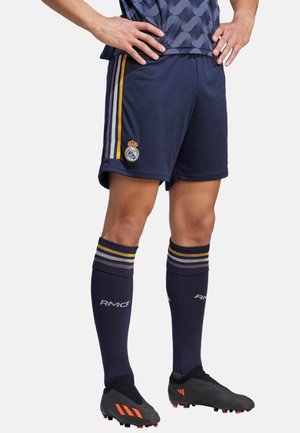 Pantalones cortos de fútbol en color marino con franjas multicolores a los lados y un escudo real; calcetas de fútbol negras con franjas; zapatos de fútbol negros con acentos naranjas.
