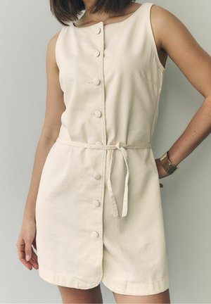 Femme portant une robe beige sans manches boutonnée avec une ceinture à la taille, debout avec une main sur la hanche et l'autre détendue vers le bas.