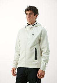 Hellbeige Kapuzenjacke mit Reißverschluss, ausgestattet mit schwarzer Hardware, einer Kordelzugkapuze und seitlichen Taschen mit Reißverschlüssen. Stoff aus Baumwollmischung mit glatter Textur.