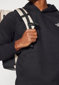 Sudadera negra con bolsillo delantero, llevada con una correa de mochila a rayas beige y negra. La mochila tiene una tela texturizada y una cremallera.