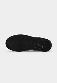 Semelle de chaussure noire avec motif de bande de roulement ondulé abstrait et forme distinctive près du talon sur un fond clair uni.
