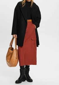 Schwarzer übergroßer Mantel, geripptes schwarzes Oberteil, rostfarbene Midirock mit Taschen, schwarze Schnürstiefel und eine beigefarbene, lässige Handtasche.