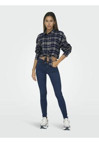 Camicia in plaid navy a maniche corte con bottoni e nodo frontale, abbinata a jeans skinny blu scuro e sneakers bianche con accenti grigi.