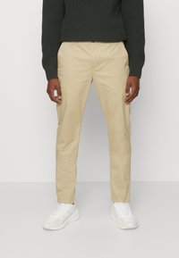 Beige Baumwoll-Chinos mit geradem Bein, seitlichen Taschen, kombiniert mit weißen Sneakern und einem dunkelgrünen Strickpullover.