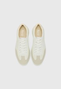 White and beige sneakers made of smooth leather and textile; features perforated detailing on the toe, flat laces, and a cushioned tongue.