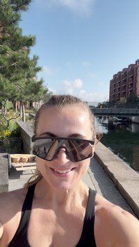 Šypsena moteris dėvi didelius akinius nuo saulės ir juodą tank topą, stovi saulėtoje taku prie kanalo, aplinkui pastatai ir žalia augmenija.
