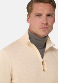 Pull-over en tricot beige avec un col zippé mi-haut et un col roulé gris clair en dessous. Présente un tirette de zip jaune et une texture côtelée.
