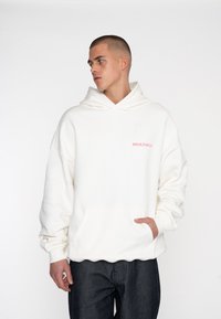 Sudadera blanca de tejido suave con un bolsillo tipo canguro, que presenta un logotipo rosa de "MULTIPLY" en el pecho y puños acanalados. Lleva puesta con jeans oscuros.