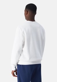 Hvid sweatshirt med rund hals og ribbede manchetter. Blød tekstur, minimalistisk design. Sættes sammen med marineblå bukser; set fra ryggen.