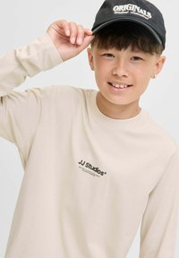 Junge lächelt, trägt ein beige Langarmshirt mit dem Text "JJ Studios" und eine schwarze Mütze mit dem Text "ORIGINALS", während er den Schirm der Mütze hält.
