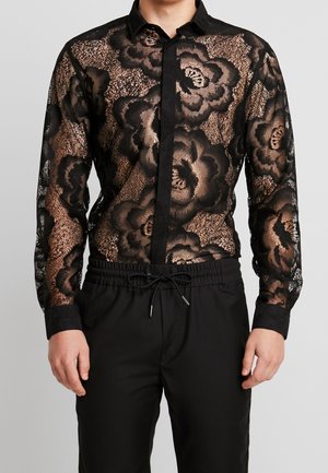 Mann trägt schwarzen, transparenten Spitzenlangarmshirt mit großen Blumenmustern und schwarze Kordelhose vor einfachem Hintergrund.