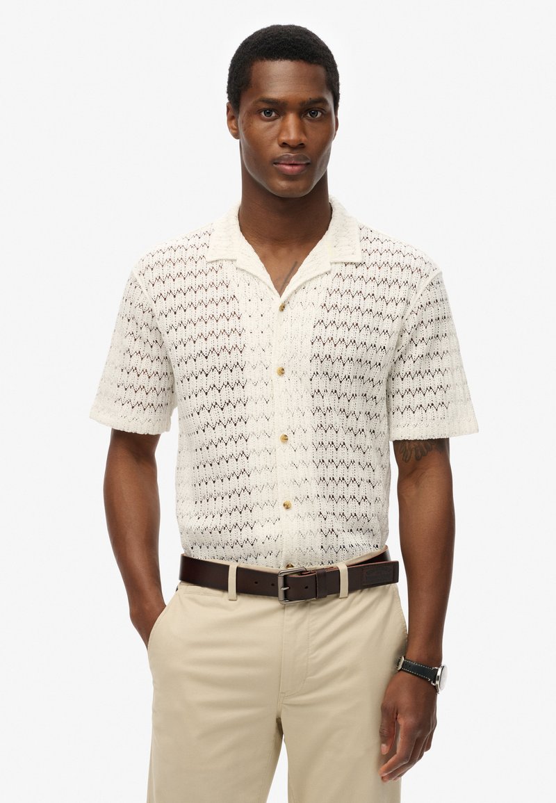 Camisa de manga corta color crema con un patrón de zigzag texturizado y cuello con muesca, decorada con pequeños botones redondos.
