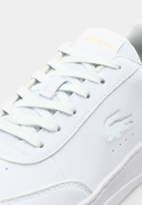 Gros plan d'une sneaker en cuir blanc avec des lacets blancs, logo crocodile en relief sur le côté, et nom de la marque sur la languette.