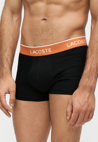 Buste masculin portant un boxer noir avec une ceinture orange de la marque "Lacoste,", montrant une partie des bras et des cuisses supérieures.