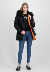 Crna parkas jakna s umjetnim krznom na kapuljači, narančastim podstavom, patentnim zatvaračima i dvostrukim gumbima. Nošena preko crnog džempera i plavih traperica s tenisicama.