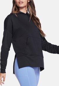 Schwarzer Kapuzenpullover mit einer Vordertasche, langen Ärmeln, seitlichen Schlitzen und lockerer Passform, kombiniert mit hellblauen Leggings.