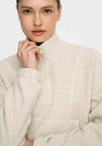 Cream-colored fleece pullover with a high collar and half zipper. It features a textured surface and fitted cuffs.