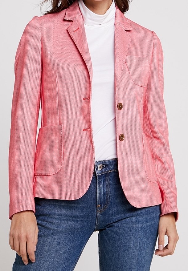 Femme portant un blazer rose cintré avec deux boutons sur un col roulé blanc et un jean bleu, debout devant un fond uni.