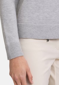 Grauer Pullover mit gerippten Bündchen und Saum, aus weichem Material. Verfügt über einen kleinen metallischen Akzent am Saum. Kombiniert mit hellen Hosen.