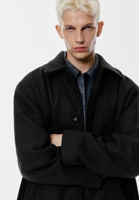 Zwarte wollen jas met een grote kraag, knoopsluiting en brede schouders. Draagt over een marineblauw overhemd met knopen. Effen achtergrond.