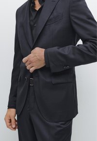 Dunkler gestreifter Anzug mit maßgeschneiderter Passform, mit spitzen Revers und Einstecktuch, getragen über einem schwarz geknöpften Hemd. Glatte Textur.