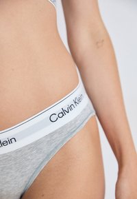 Close-up van de taille van een persoon die grijze Calvin Klein onderbroeken draagt met een witte gebrandmerkte tailleband, met een gedeeltelijk zichtbare arm en torso.