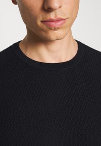 Suéter de punto negro texturizado con cuello redondo; vista en primer plano que enfatiza los detalles de la tela y el diseño del cuello.
