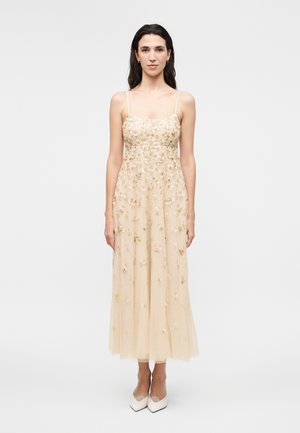 Žena stojí čelom vpredu, oblečená v bezrukávové béžové midi šaty s kvetinovou výšivkou a bielymi lodičkami so špicatou špičkou na jednoduchom pozadí.