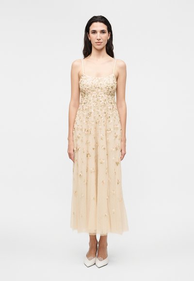 Woman stands facing forward wearing a sleeveless beige midi dress with floral embroidery and white pointed-toe heels on a plain background.
