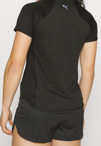 Černé sportovní tričko s krátkými rukávy, textilní materiál a malé fialové logo na zádech. V kombinaci s černými šortkami s bočními rozparky.