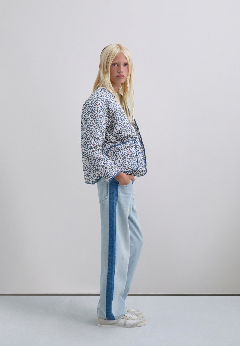 Chaqueta acolchada con un patrón floral azul, combinada con jeans de un azul claro que tienen una franja lateral de un azul oscuro. Zapatillas blancas completan el conjunto.
