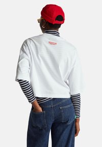 Bílý krátký top s červeným logem "Stranger Things" na zádech, nošený přes námořnicky modrý a bílý proužkovaný dlouhý rukáv, doplněný modrými džíny.