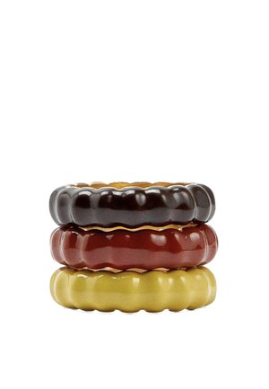 Stos trzech błyszczących, żebrowanych ceramicznych pierścieni w kolorze czarnym, głębokiej czerwieni i musztardowożółtym na białym tle.