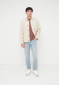 Μπεζ βαμβακερό μπουφάν με κουμπιά, δύο τσέπες στο στήθος, φορεμένο πάνω από ένα καφέ τοπ, συνδυασμένο με ανοιχτό μπλε τζιν και λευκά αθλητικά παπούτσια.