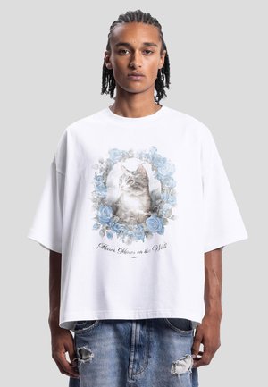 Persona con capelli intrecciati che indossa una maglietta bianca oversize con un ritratto di gatto circondato da rose blu e testo, abbinata a jeans strappati.