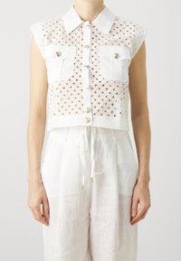 Krótkie białe koszuli z bezrękawnikowym fasonem, z siatkowym wzorem, dwoma kieszeniami na piersi oraz dekoracyjnymi złotymi guzikami. Detal z fakturowanej tkaniny.