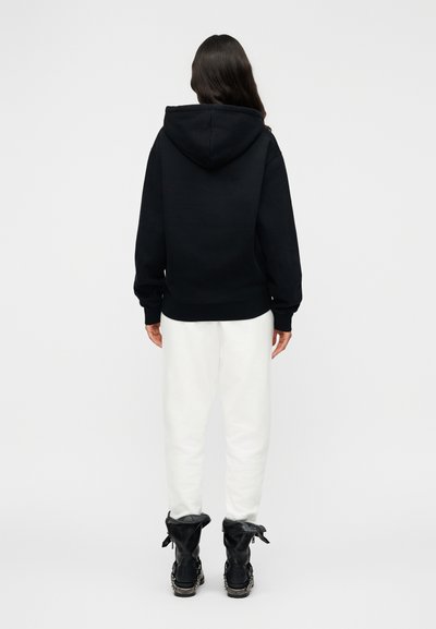 Sweat-shirt à capuche noir avec une coupe décontractée, associé à un pantalon de survêtement blanc et des bottes noires à lacets ornées d'un détail chaîne décoratif.