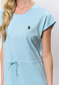 Rochie albastru deschis cu mâneci scurte, realizată dintr-un material moale, prevăzută cu o talie ajustabilă prin șnur și un mic logo brodat pe piept.