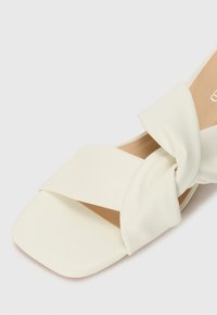 Sandale à enfiler en cuir synthétique blanc avec un design de strap noué et un bout carré. Texture douce et lisse, avec une semelle intérieure beige.