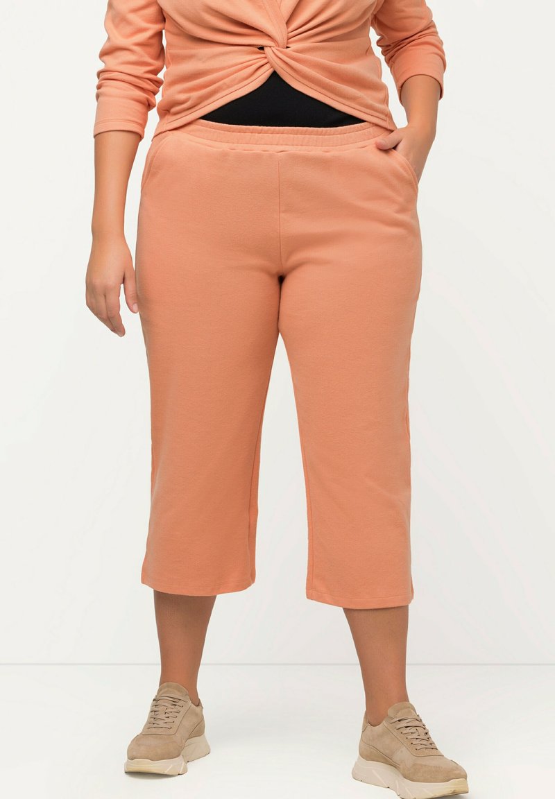 Kvinde iført ferskenfarvede cropped sweatpants, ferskenfarvet vridning af langærmet top og beige sneakers, stående med den ene hånd i lommen.