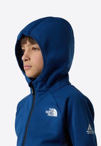 Kék kapucnis dzseki puha anyagból, elöl cipzárral, a mellrészen a The North Face logóval, az ujján pedig a Mountain Athletics logóval.