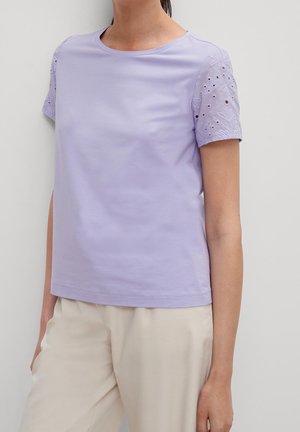 Mujer con una blusa de manga corta lila con bordado floral de ojales en las mangas y pantalones beige, sobre un fondo liso.