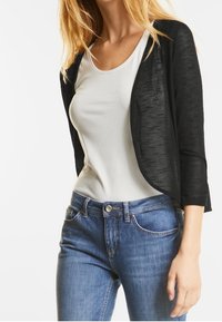 Svart, lätt, kort cardigan över en vit topp, kombinerad med blå jeans. Cardiganen har en slät textur och öppen framdesign.