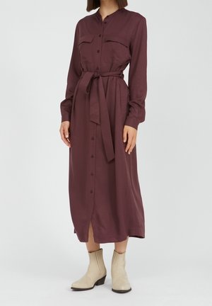 Kobieta w bordowej sukience z długim rękawem, zapinanej na guziki, z paskiem, w połączeniu z jasnobeżowymi botkami na płaskim obcasie na jednolitym tle.